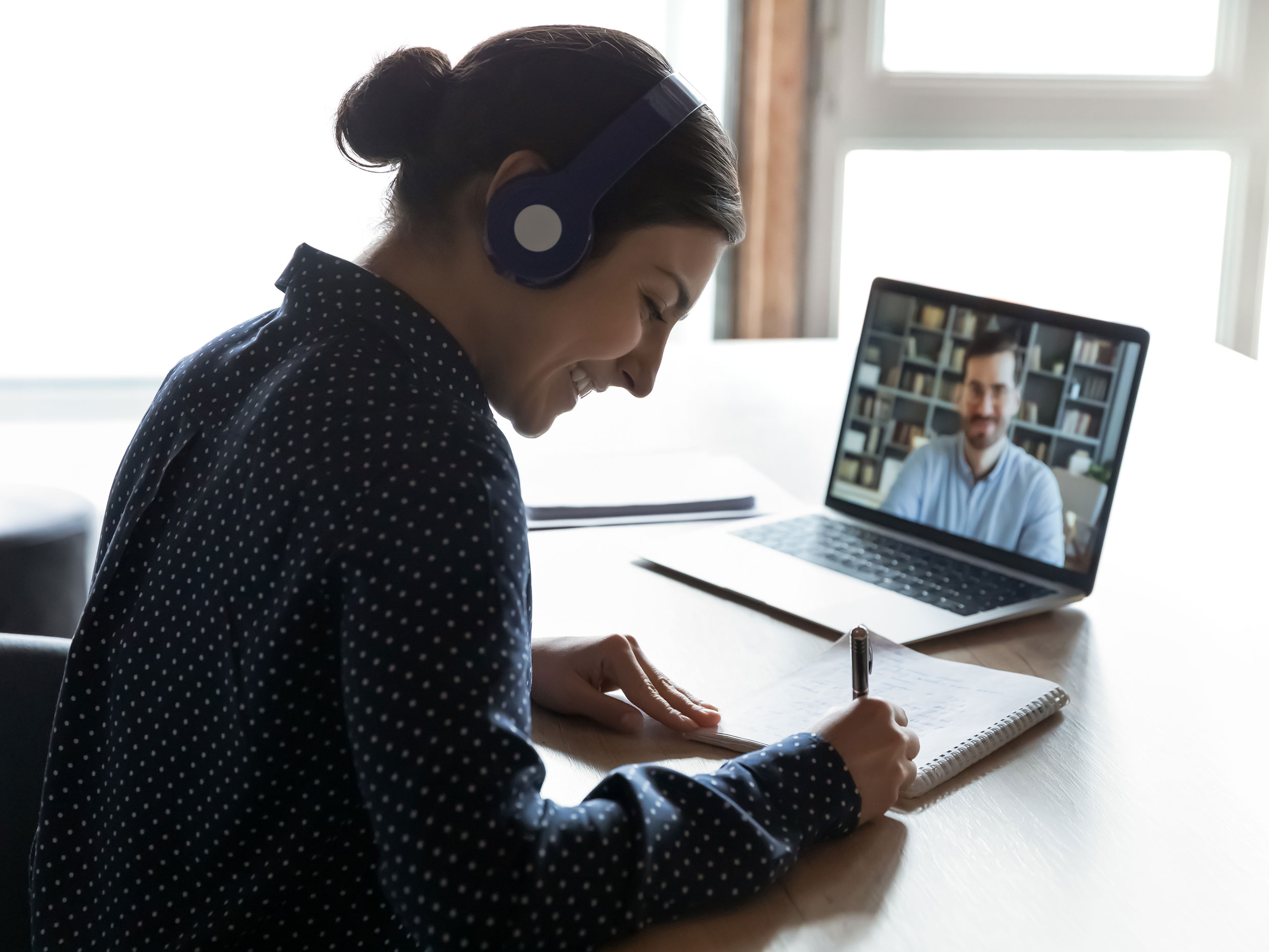 A smiling female student sitting in front of her laptop with headphones on her head having an online class with her instructor and making some notes.
