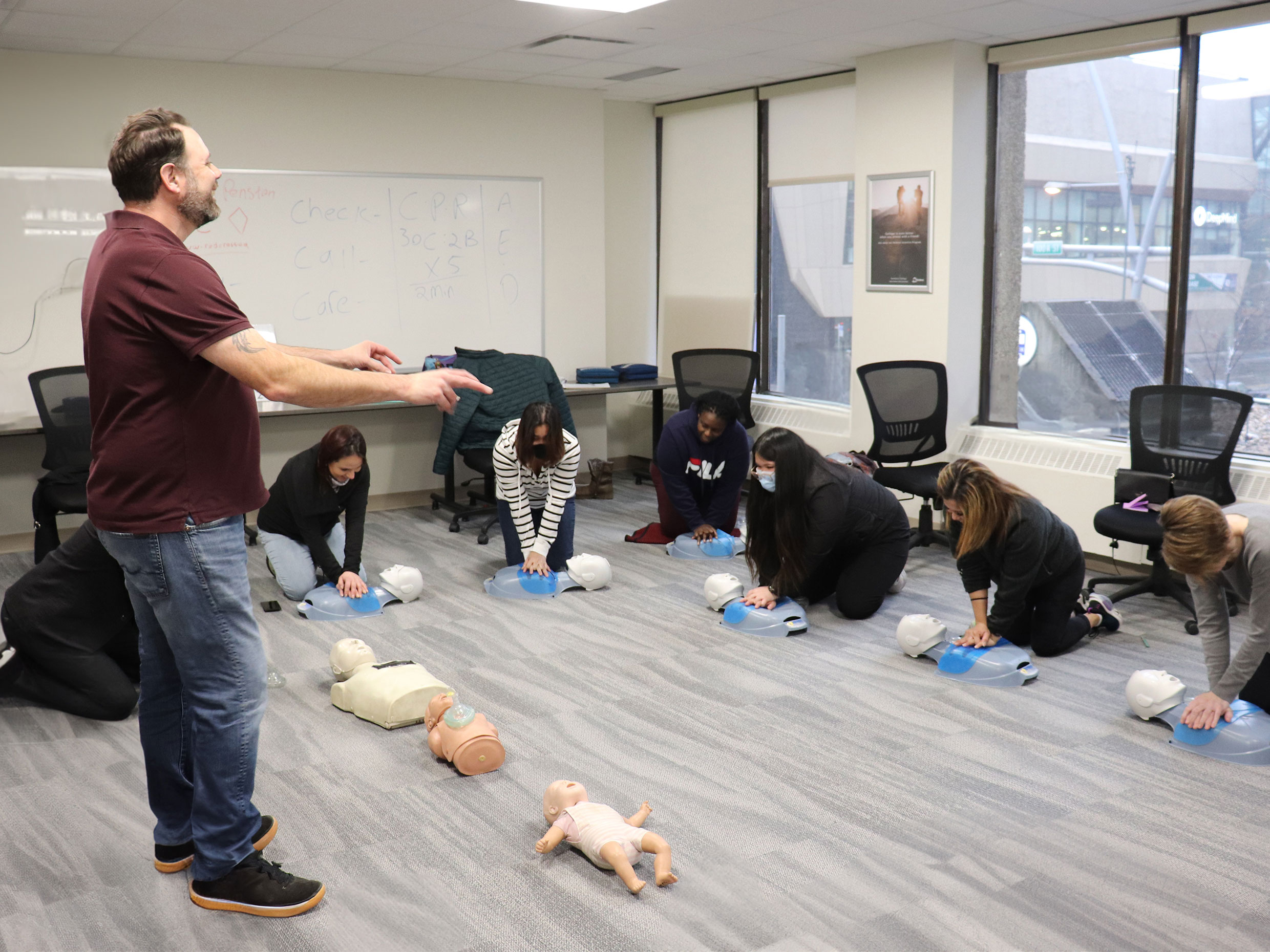 The CRP and First Aid course instructor standing in the middle of a classroom surrounded by students learning how to administer first aid on models on Sundance College's Edmonton campus.
