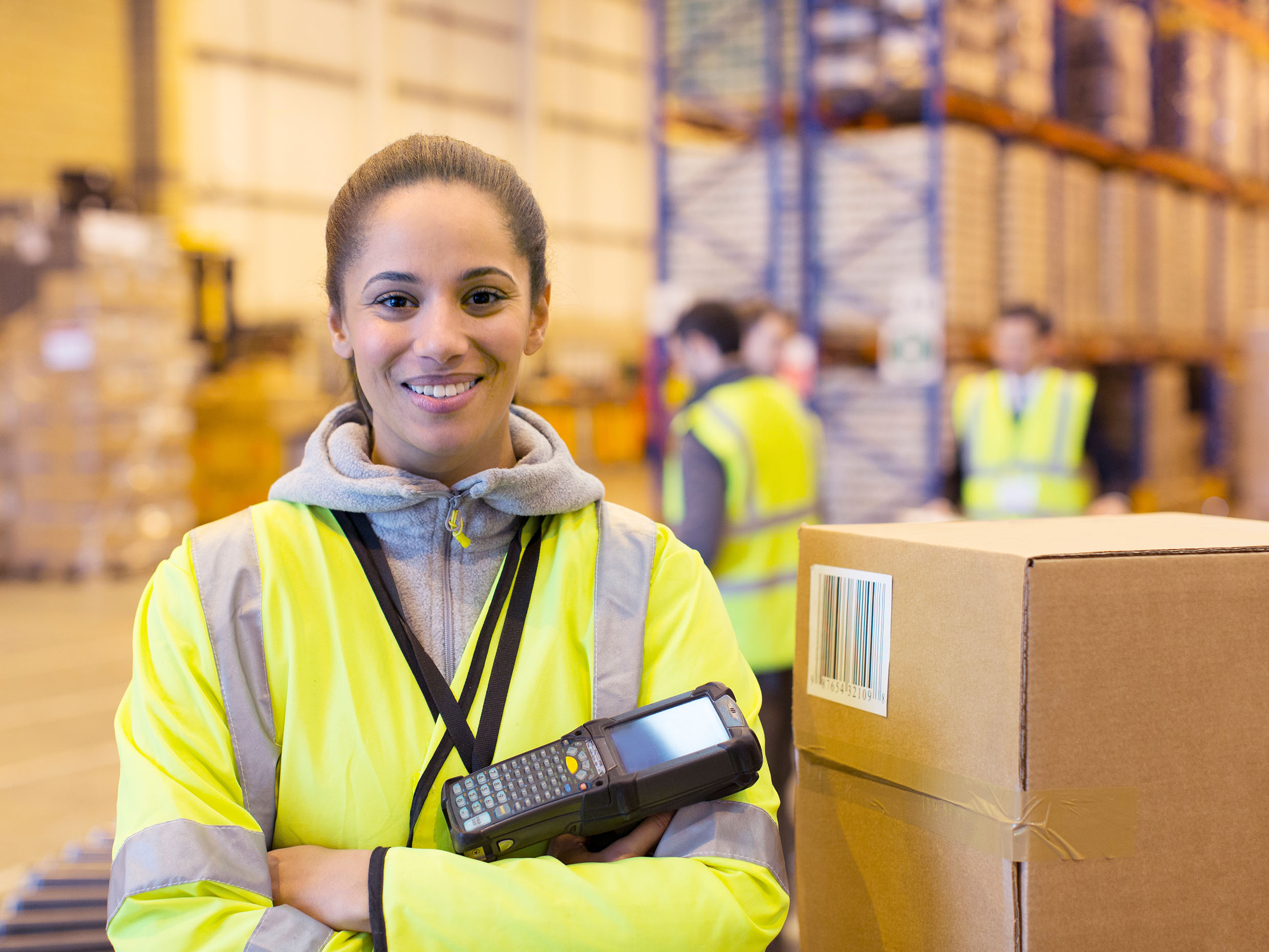 A happy logistics coordinator female standing in a warehouse and holding a pos terminal device in her hands.