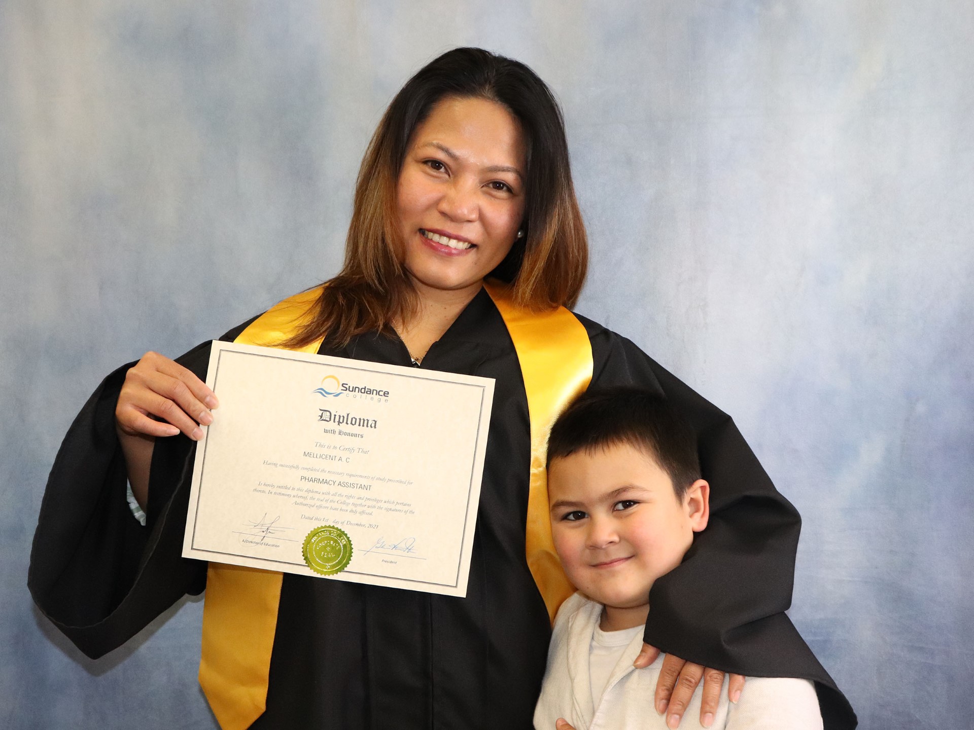 A happy Sundance College graduate holds her Pharmacy Assistant Diploma in one hand as she cuddles her son in the other.