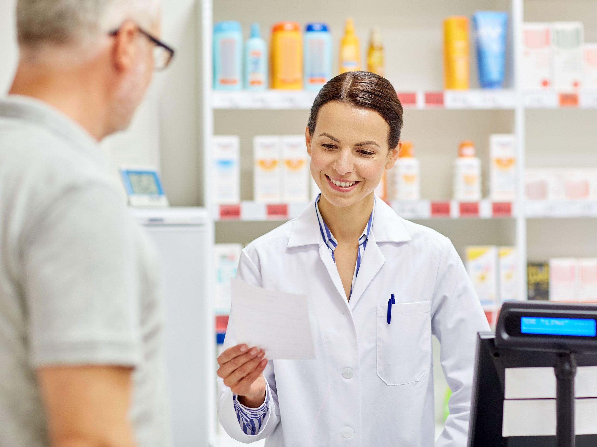 Woman in lab coat standing behind pharmacy counter and holding a piece of paper, with customer facing her from the other side of the counter.