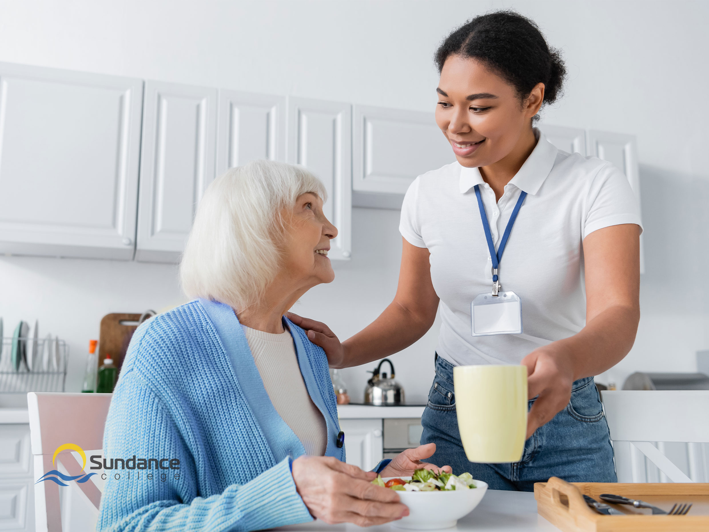 Young female personal support care worker in the kitchen with an old lady giving her a cup of tea or coffee as she eats a salad lunch