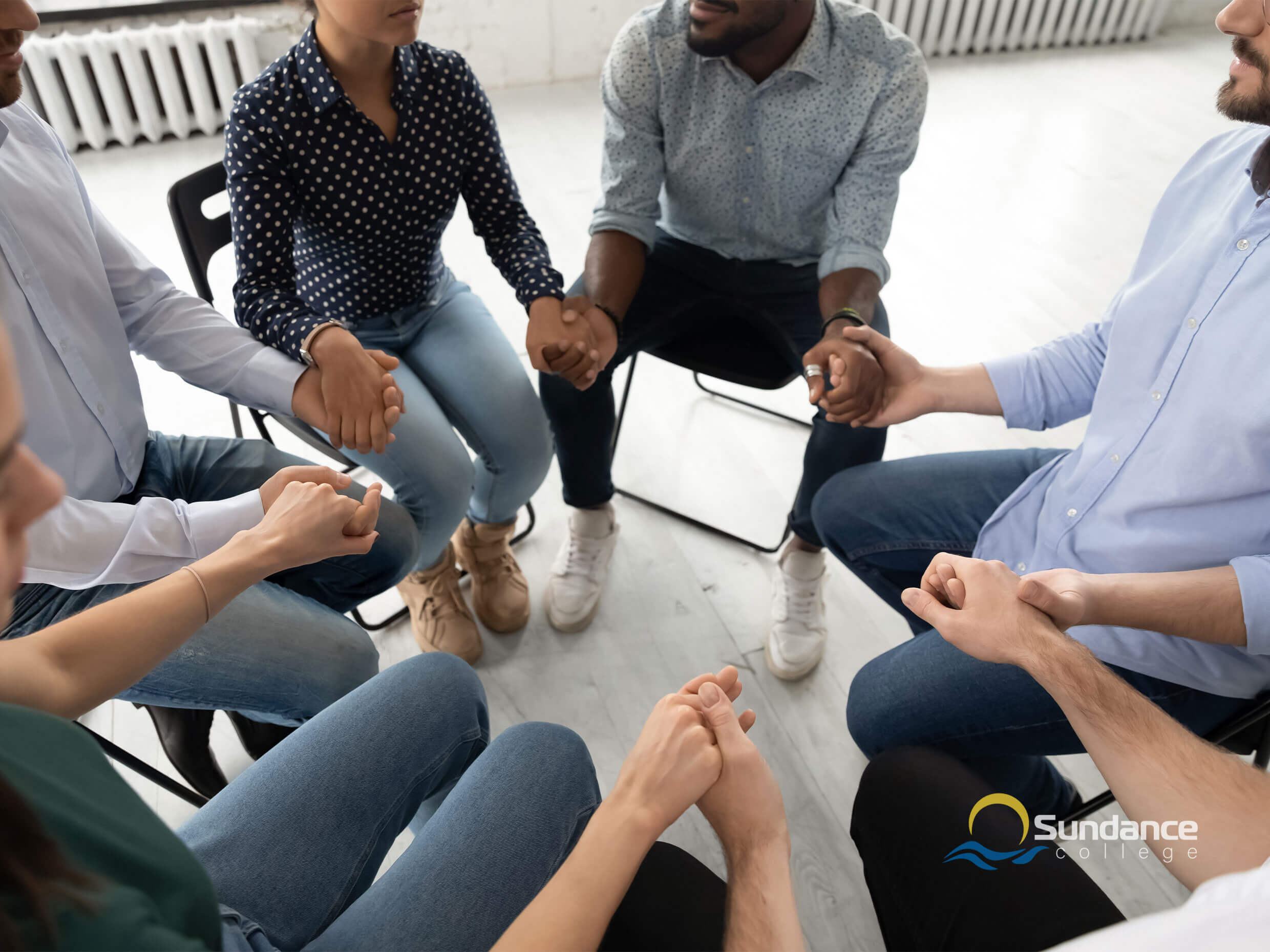 A circle of people in chairs holding hands as part of a group therapy session run by an addictions worker trained in evidenced-based community health procedures