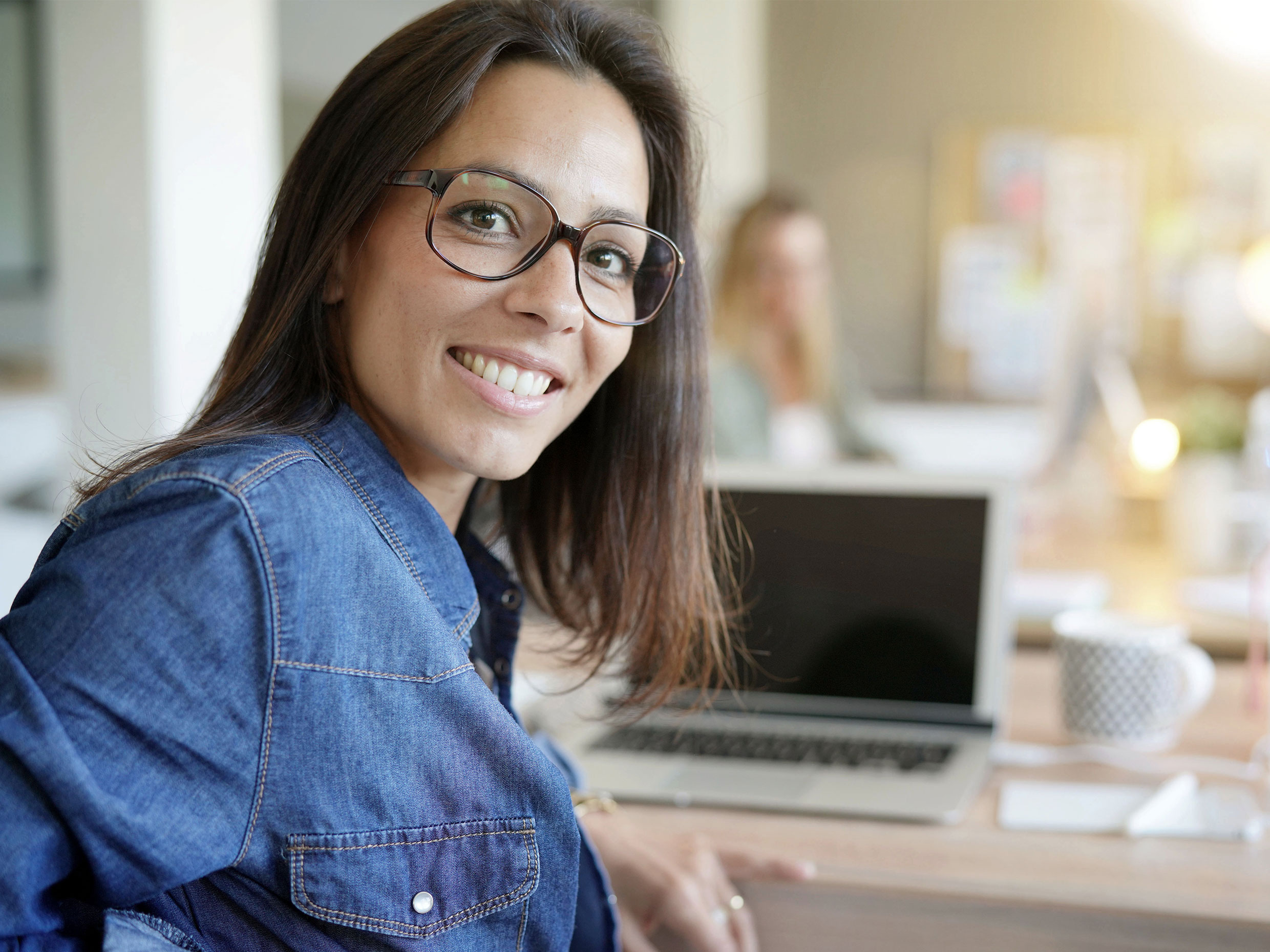 40-something woman in glasses smiling while looking over shoulder with laptop in background