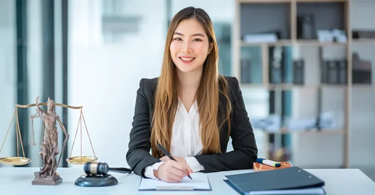 Smiling woman in business attire writing on a clipboard at a desk with gavel, scales, and legal books in a law office setting.