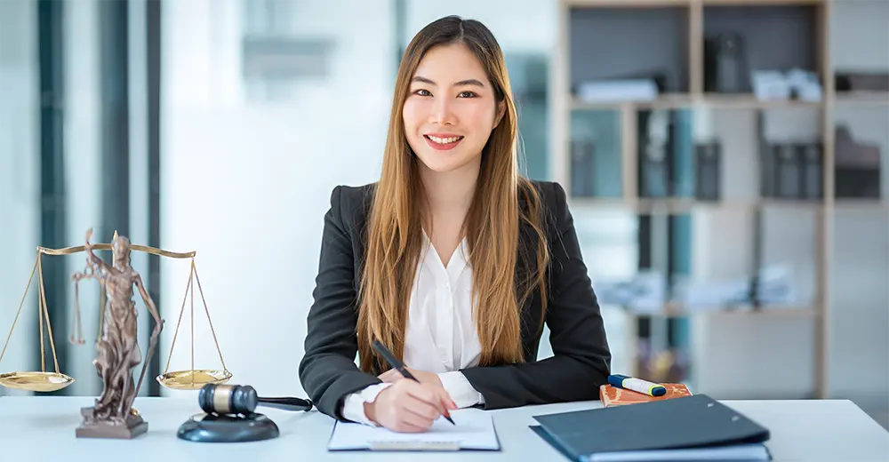 Smiling woman in business attire writing on a clipboard at a desk with gavel, scales, and legal books in a law office setting.