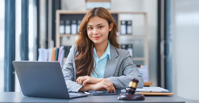 Smiling legal assistant in a blazer holding a clipboard at a desk, with legal scales, a gavel, and office files in the background.