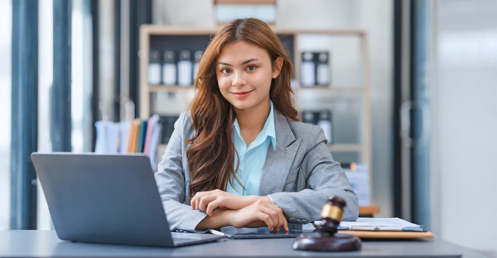 Smiling legal assistant in a blazer holding a clipboard at a desk, with legal scales, a gavel, and office files in the background.