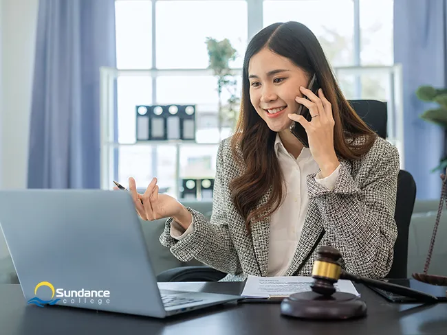 Smiling legal assistant reviewing paperwork at a desk with Lady Justice statue, legal scales, gavel, and books in view.