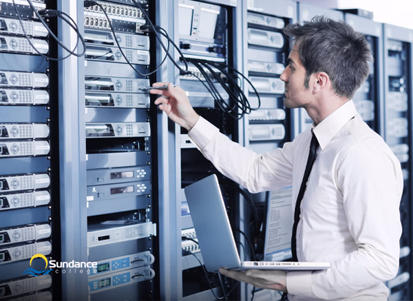 A Network Systems Manager holds a laptop in one hand as he examines cables on a server rack.