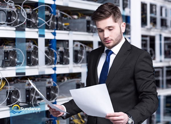 A Network Systems Manager holds a laptop in one hand as he examines cables on a server rack.