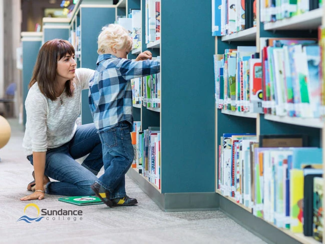 Education assistant helping young student pick out a book in the classroom bookshelf.