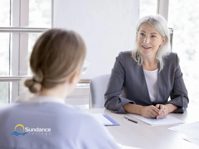 In a sunlit office, a prospective student of Sundance College engages in a conversation with her employer, who dons a sleek grey jacket, discussing her educational and professional aspirations.