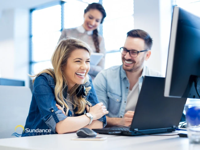 Three Sundance College Mobile and Web Developer Diploma program graduates beam with happiness as they receive their first paychecks from their employer. They're seated at a desk in a well-lit office, exuding satisfaction.