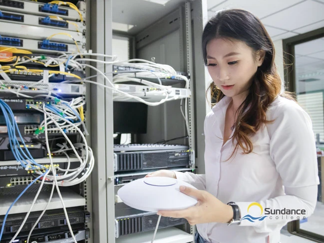 A Network Systems Manager Diploma graduate from Sundance College poses confidently in front of server racks, showcasing diverse cable networks. She holds a Wi-Fi router with a glowing green indicator, symbolizing proficiency in network management.