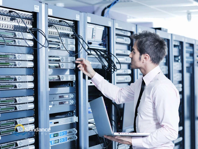 A Network Systems Manager Diploma holder from Sundance College stands before server blocks, attentively monitoring cable connections and sockets. With a laptop in hand, he demonstrates expertise in network monitoring and server administration.