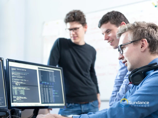 Sundance College Mobile and Web Developer Diploma program graduates collaborate at a software development company. They're seen coding a website on their computer screens while discussing project functionality.