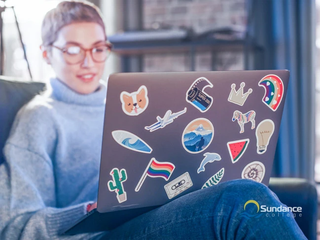 A web developer who graduated from Sundance College is sitting on the couch, working on her laptop, adorned with lots of funny and colorful stickers.