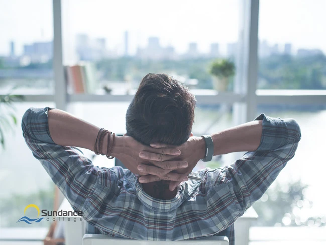 A student from Sundance College relaxes in a chair, hands crossed behind his neck, gazing out of a window. Outside, he sees lush green trees, colorful flowers, towering skyscrapers of a big city, and a bright blue sky, taking a break from his online learning.