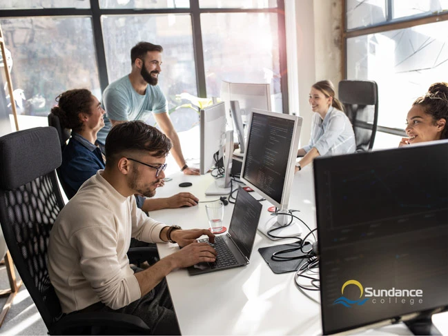 A mobile and web developer graduated from Sundance College sitting in front of his computer in the office and analyzing the lines of code surrounded by other team members.