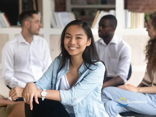 A young woman sits facing the back of her chair in the foreground, while a group of individuals is seated in the background for a mental health support group session.