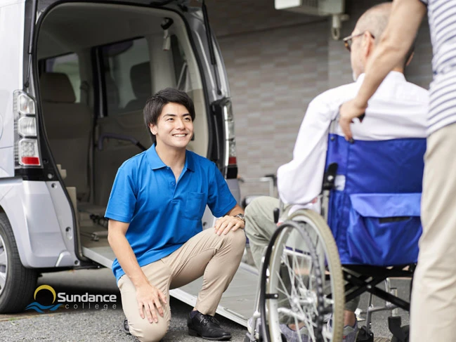 Young male personal support care worker in blue scrubs tucking an old lady into bed in her home or nursing home