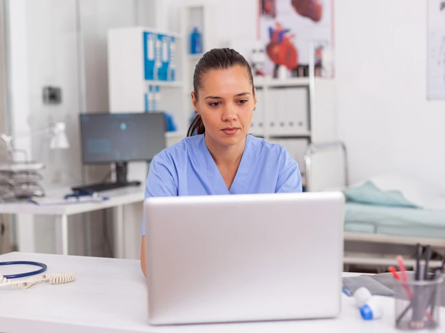 a woman in a blue scrubs sitting at a desk with a laptop