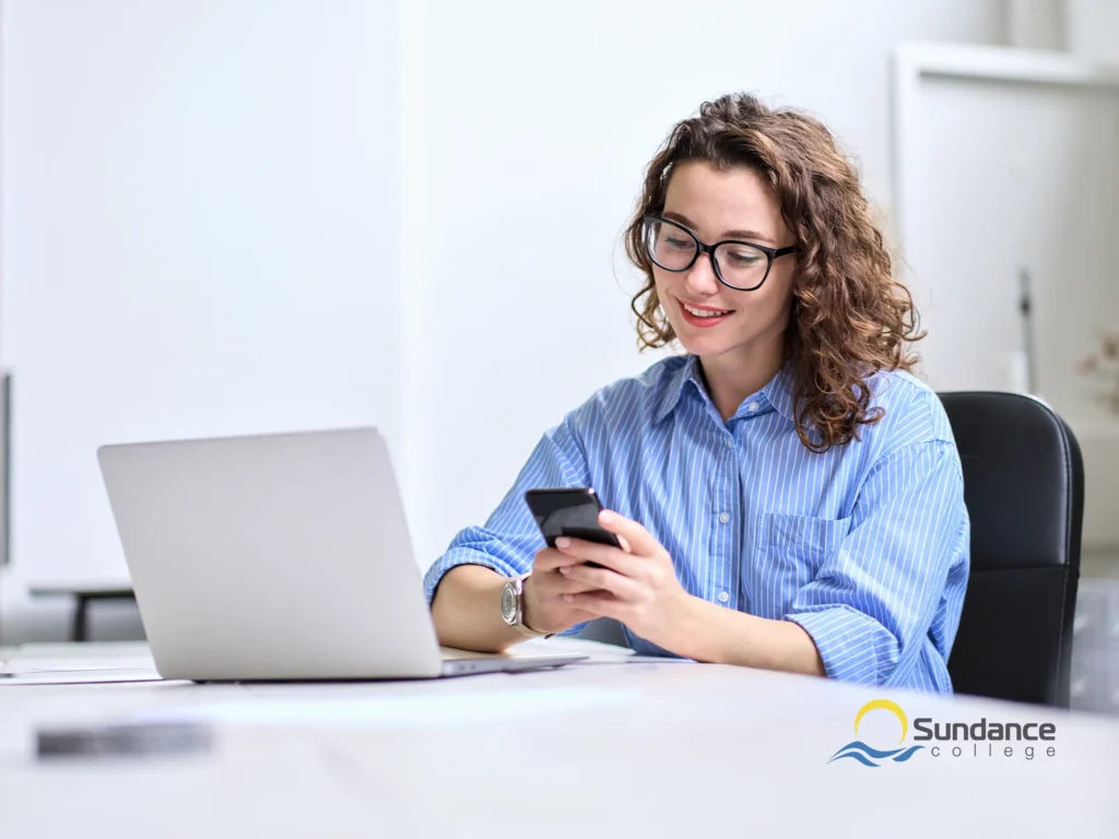 Sundance College Mobile and Web Developer Diploma graduate sitting at her office desk, holding a mobile phone and smiling while learning new cross-platform app development frameworks on her laptop.