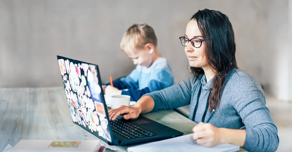 An adult student mom learning online, taking notes while looking at her laptop screen, with her young son drawing beside her.