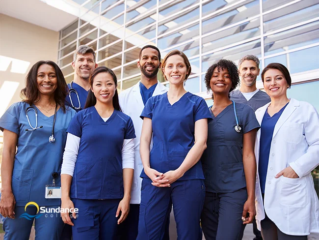 Numerous smiling personal support workers and other healthcare professionals stand in front of a medical setting.