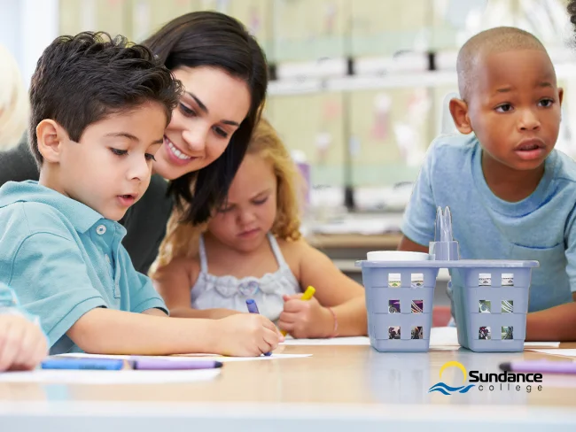 A smiling education assistant observes as several children draw with crayons in a bright, well-lit classroom.