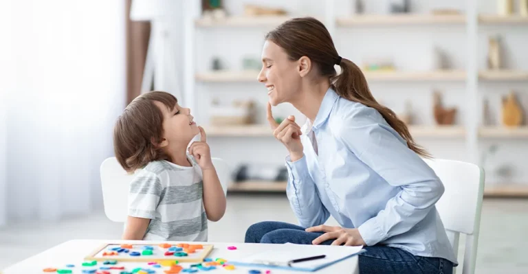 A cheerful educational assistant sits next to a 4-year-old boy, both playfully touching their chins while engaged in a fun learning game.