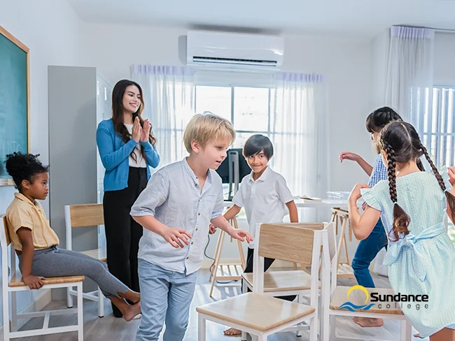 A teacher assistant claps her hands as first-grade students play a game of musical chairs, racing to sit down at the signal in a lively classroom.