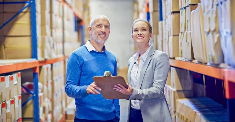 Supply chain manager in a warehouse wearing a high-visibility jacket and a helmet, looking intently at a computer.