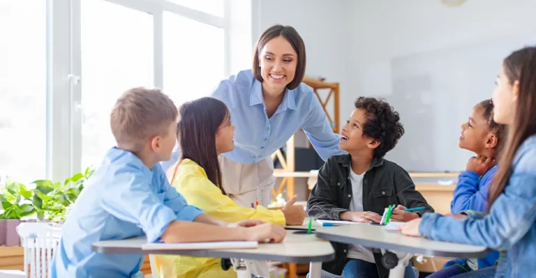 A young female education assistant talking to a group of diverse elementary students seated close to each other in a semi-circle in class.