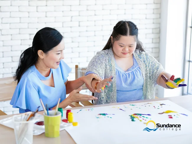 A young female special education aide showing an elementary school girl with unique learning needs how to make colorful palm imprints on a blank canvas.