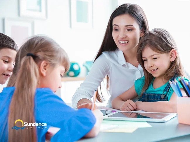 A young female homework assistant smiles, watching over three elementary students as they write in their notebooks at a shared desk in class.