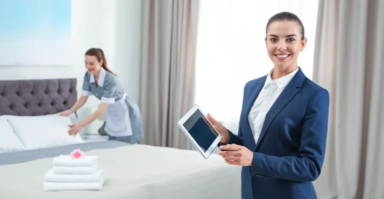 Female housekeeping supervisor holding a tablet, standing in a hotel room with a staff member cleaning in the background.