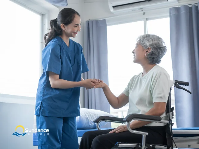 A professional caregiver gently holds the right arm of a female patient in a wheelchair, both sharing a joyful smile in a hospital room.