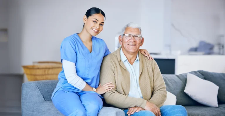 A personal support worker and an elderly man sitting together on a couch in a nursing home, both smiling warmly and enjoying each other's company.