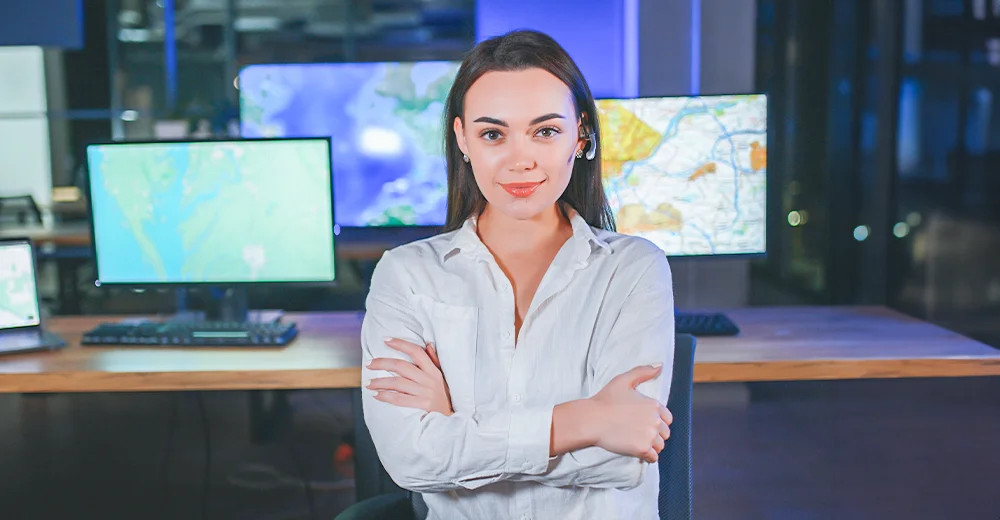 Female supply chain technician standing confidently in front of monitors displaying world maps and logistics data.