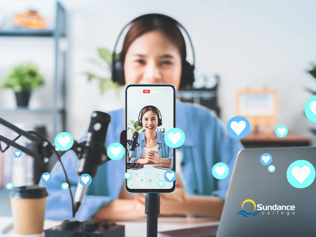 Young female social media influencer sitting at a table, live streaming with her smartphone on a stand. In front of her are a microphone and laptop, creating a professional setup for engaging with her audience.