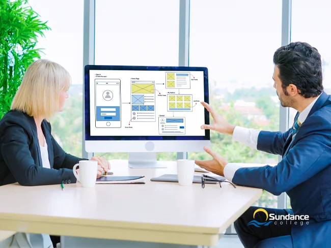 Two IT professionals seated at a shared desk, discussing a mobile app design displayed on their computer screen.