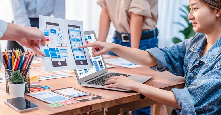 A young female app developer pointing toward a mock design on paper held by a colleague while working on her laptop at the office.