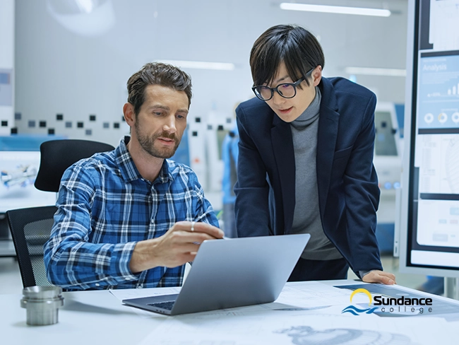 Two network professionals collaborate in an office, one seated pointing at a laptop, the other standing and observing.