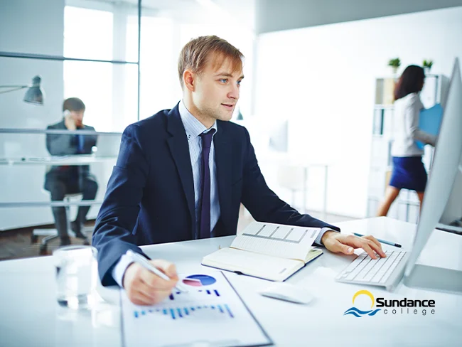 Bookkeeper working on a computer system, tracking company finances with a financial report beside them.