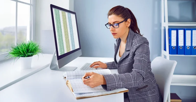 Bookkeeper noting down journal entries in a ledger while working on a computer system.