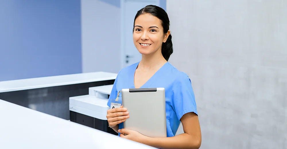 Cheerful young woman wearing blue uniform, standing at a hospital front desk with a welcoming smile. The setting features a clean, professional healthcare environment with light colors, emphasizing a friendly and approachable atmosphere.