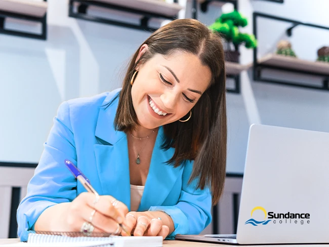 Smiling woman in an orange blazer writing her career development plan in a notebook at a desk with a laptop.