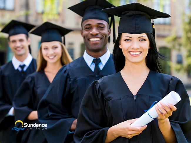 Group of smiling graduates in caps and gowns standing in a line outdoors, holding diplomas tied with blue ribbons.